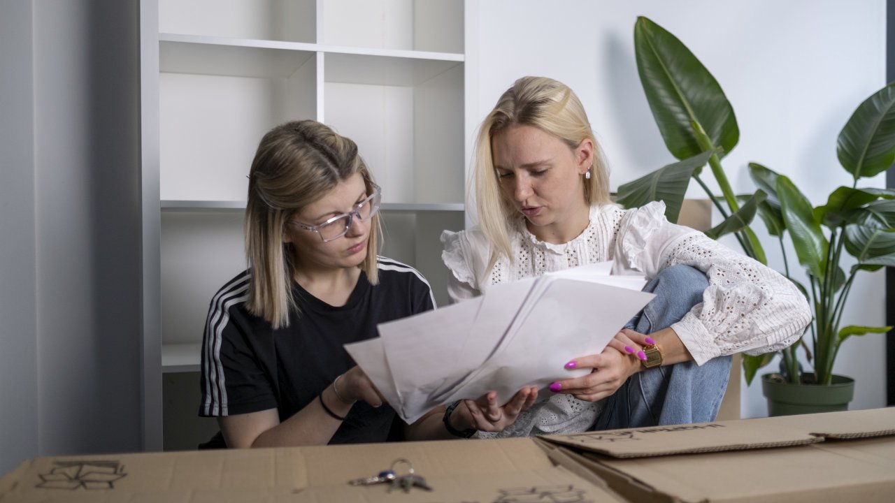 Zwei weiblich gelesene Personen, umgeben von Umzugskartons, schauen sich einen Mietvertrag an.