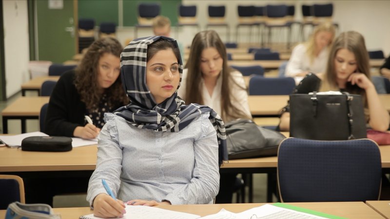 Das Bild zeigt eine junge Frau und weitere junge Menschen im Hintergrund. Sie alle sitzen in einem Klassenzimmer.